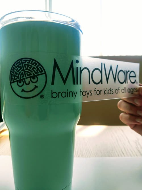 A custom clear decal reading 'MindWare: brainy toys for kids of all ages' being applied to a light teal tumbler.
